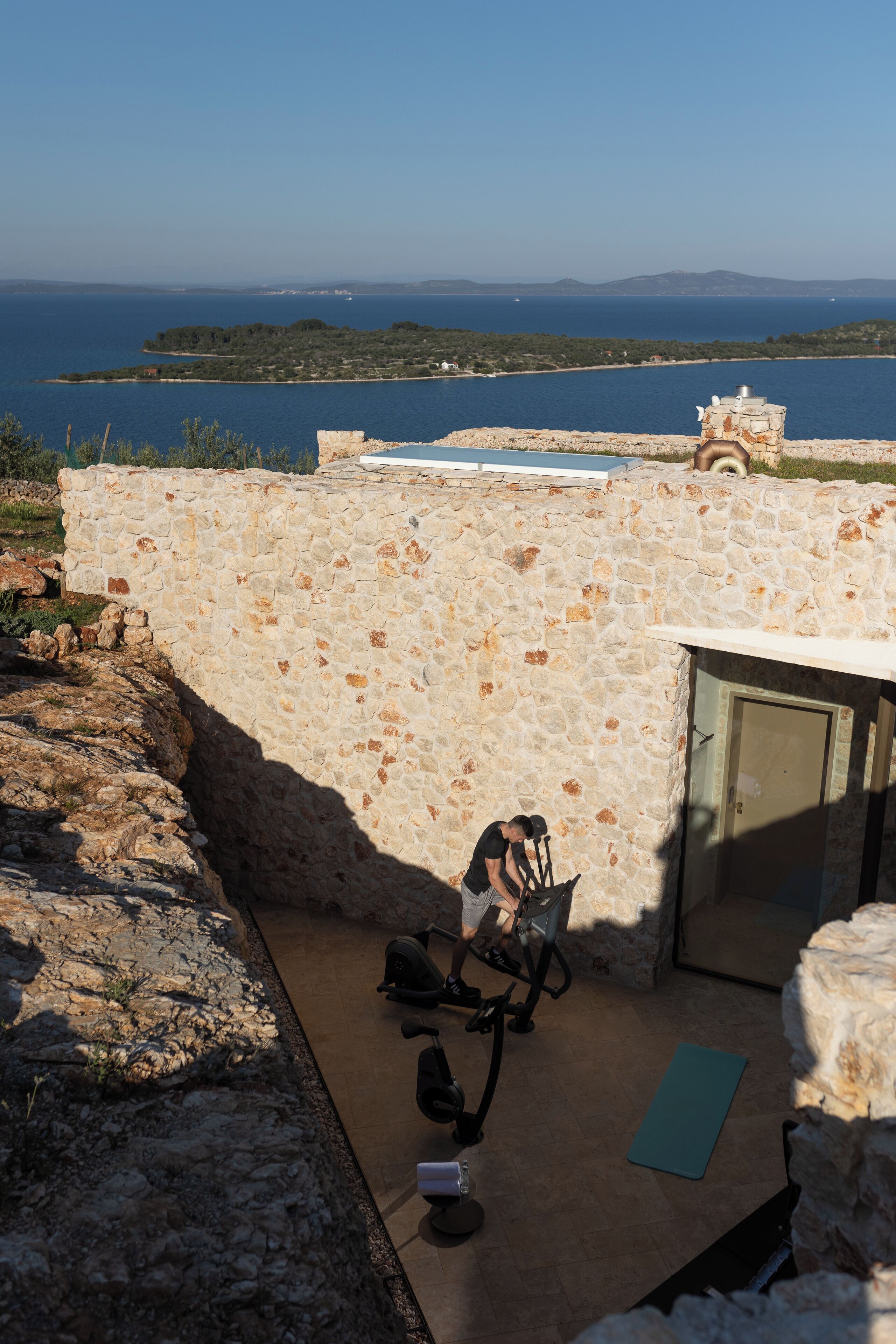 Un espace extérieur entouré de murs en pierre, équipé de matériel d’exercice, d’un tapis de yoga et offrant une vue sur la mer et les collines lointaines.