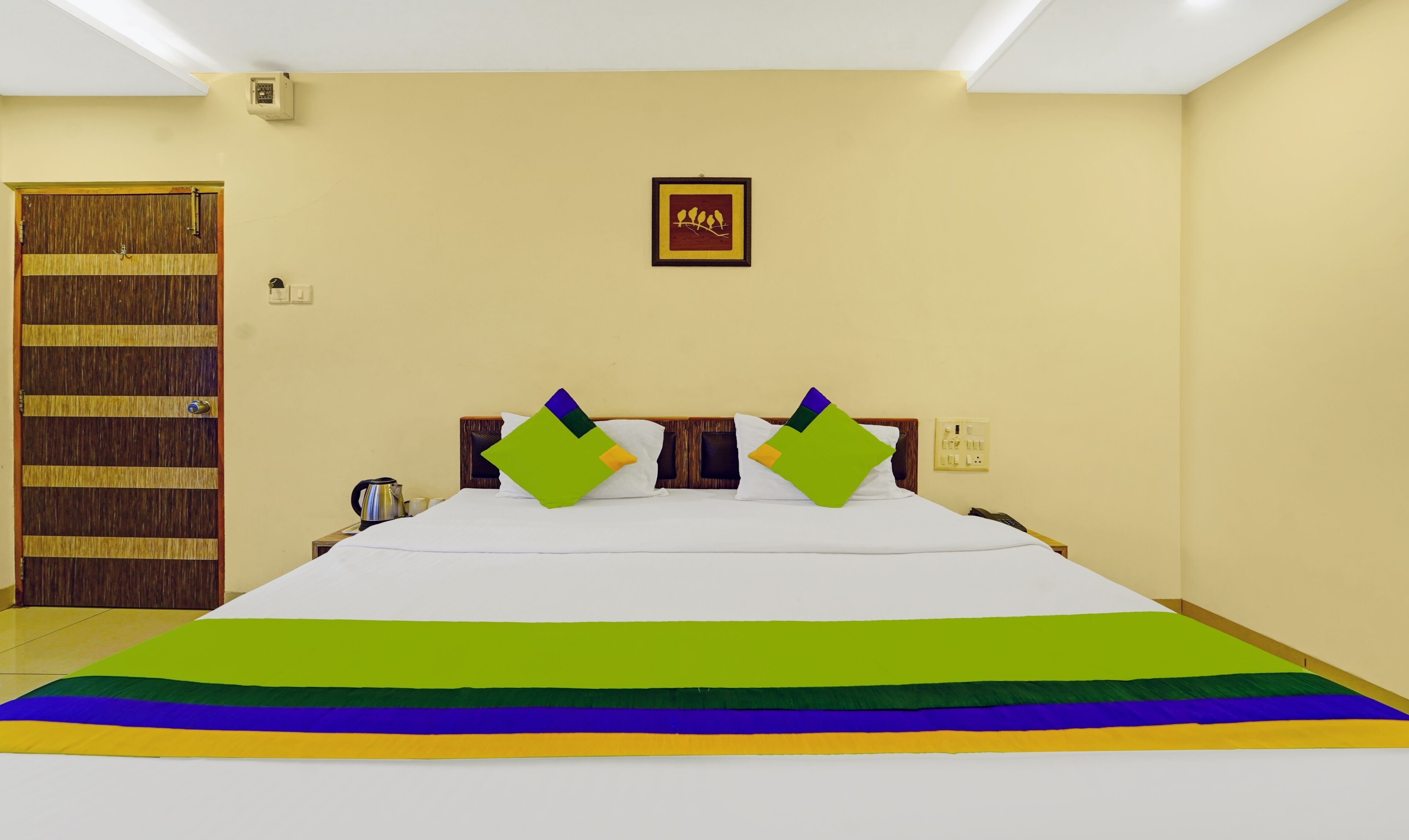 A hotel room with a large bed, a wooden door, a framed picture on the wall, and a nightstand with a kettle.