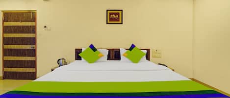 Habitación de hotel con una cama grande, una puerta de madera, un cuadro enmarcado en la pared y una mesita de noche con un hervidor.