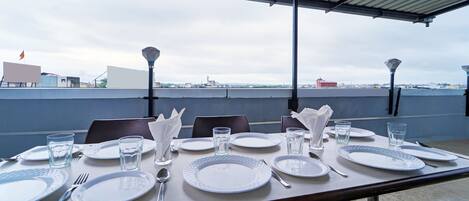 Un área de comedor en la azotea con mesas dispuestas para cuatro, cada una con platos, cubiertos y vasos. Se observa un techo metálico y una vista del paisaje urbano de fondo.