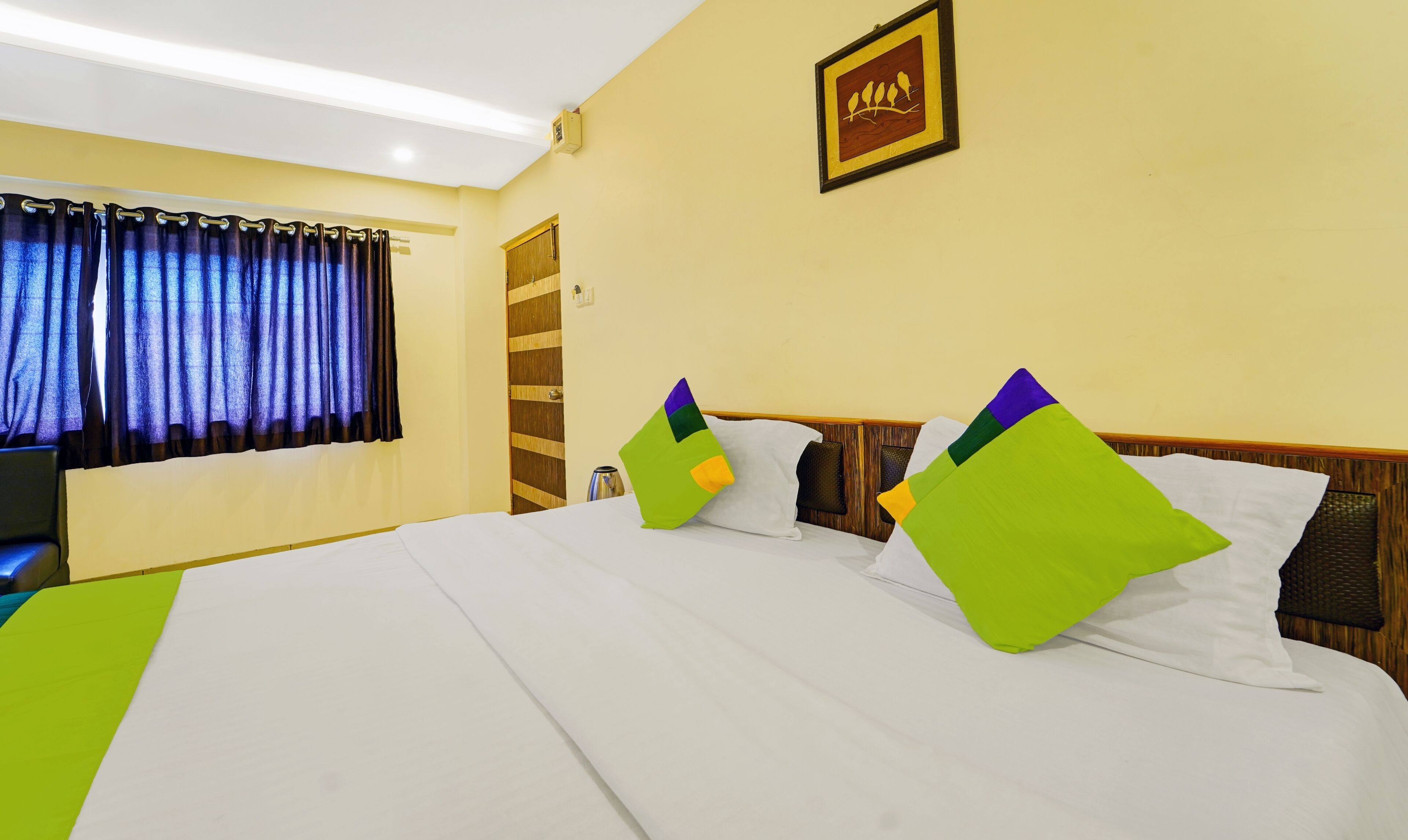 A hotel room with a large bed, two pillows, a wooden headboard, a framed picture on the wall, and a window with curtains.