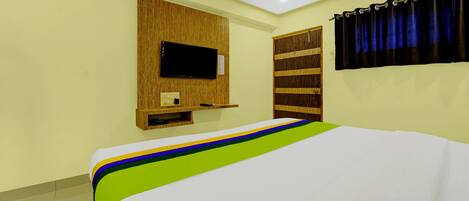 A hotel room with a bed, a TV mounted on a wooden panel, a wardrobe, and a curtain.