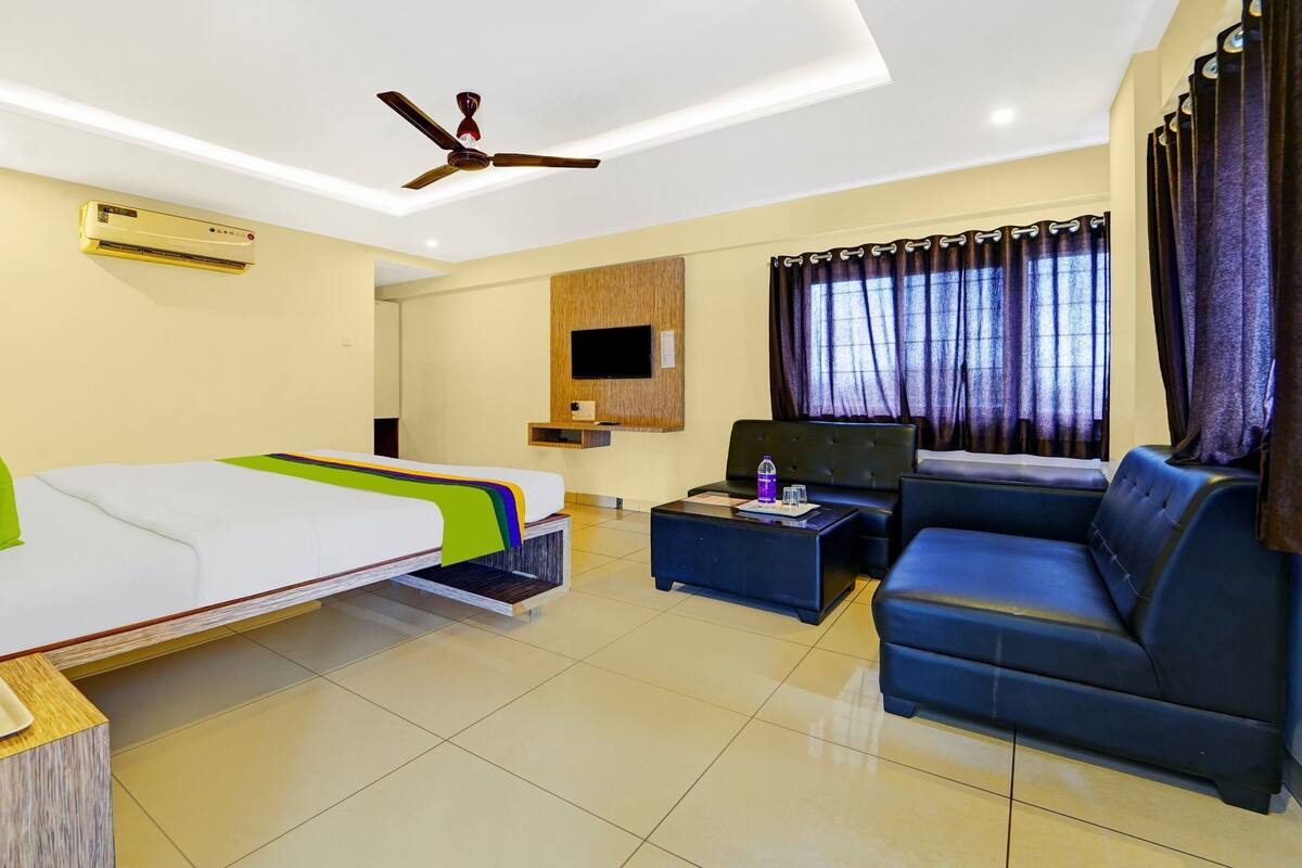 A hotel room with a bed, sofa, TV, air conditioning unit, ceiling fan, and a window with curtains.