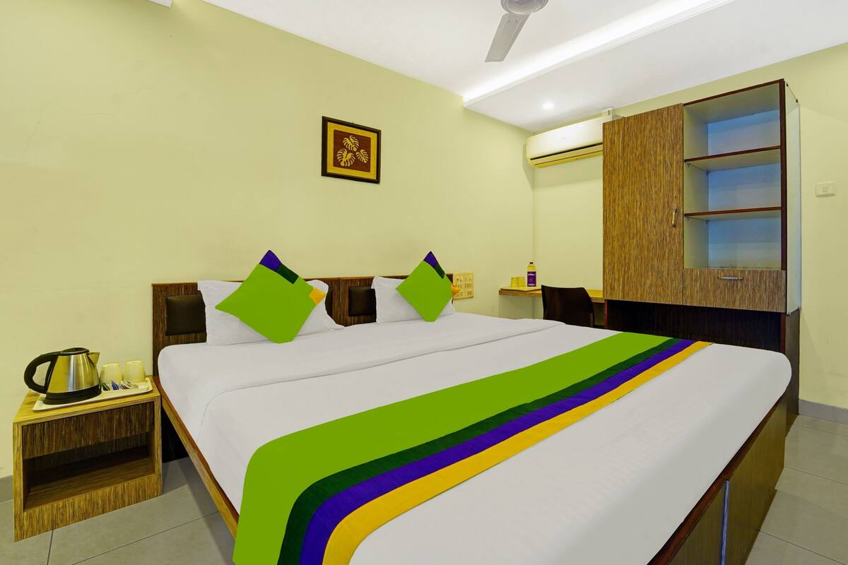 A hotel room with a large bed, a wooden nightstand, a small table with a kettle, and a wardrobe with a mirror.