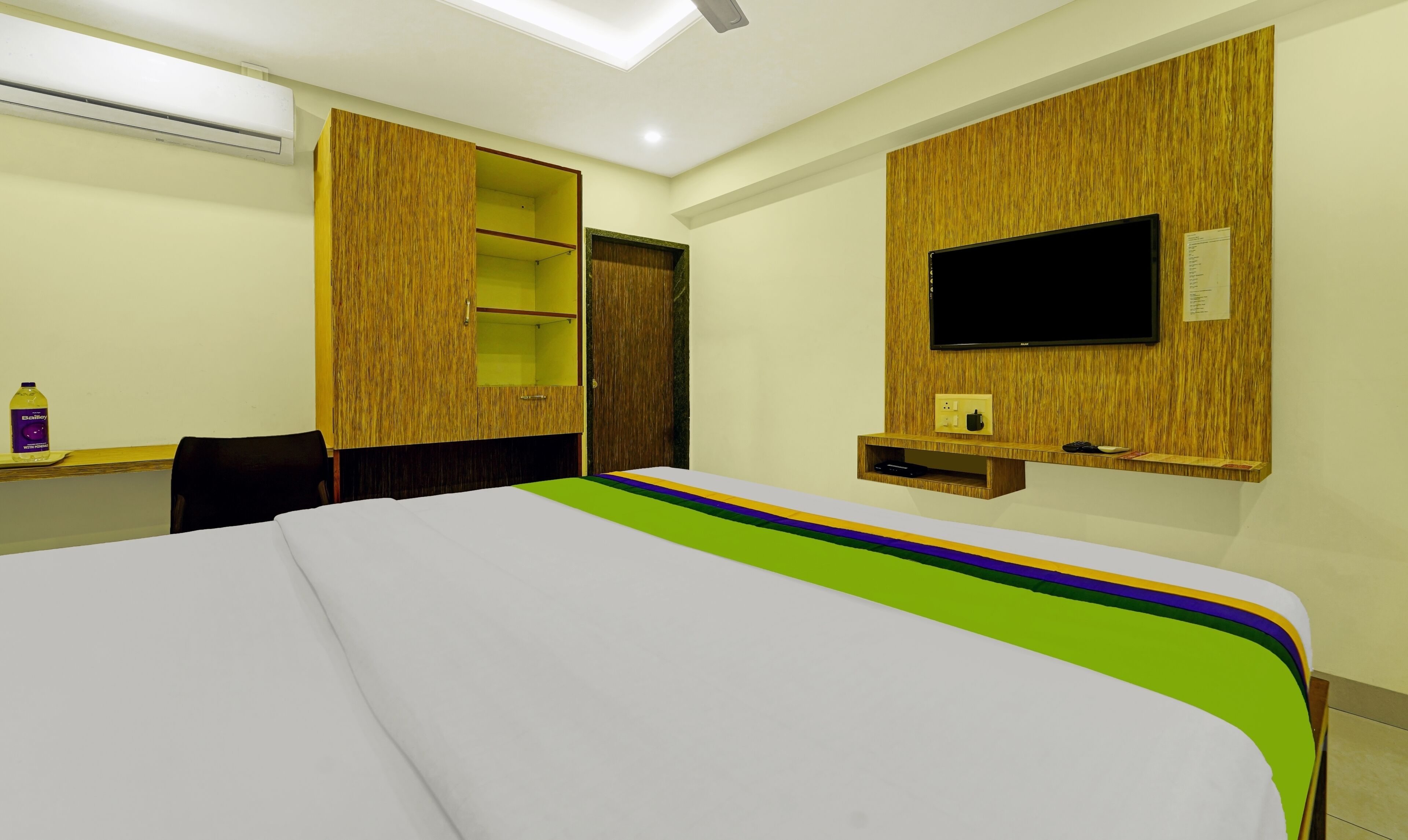A hotel room with a bed, a desk, a chair, a TV mounted on the wall, and a wardrobe with a mirror.