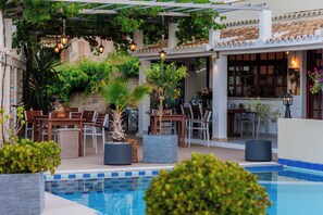 Un área junto a la piscina con una pérgola, asientos y plantas en macetas.
