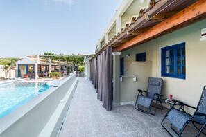 Zona junto a la piscina con vista despejada, un área de descanso cubierta con sillas y un edificio con tejado de baldosas y ventanas azules.