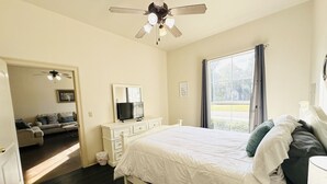 Et soveværelse med seng, ventilator, kommode, fjernsyn og et vindue med gardiner.