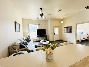A spacious living room with a sofa, a coffee table, and a TV. There is a ceiling fan and a door leading to another room. - Chateau 4, spacious townhouse in Tickfaw, LA (Tickfaw)