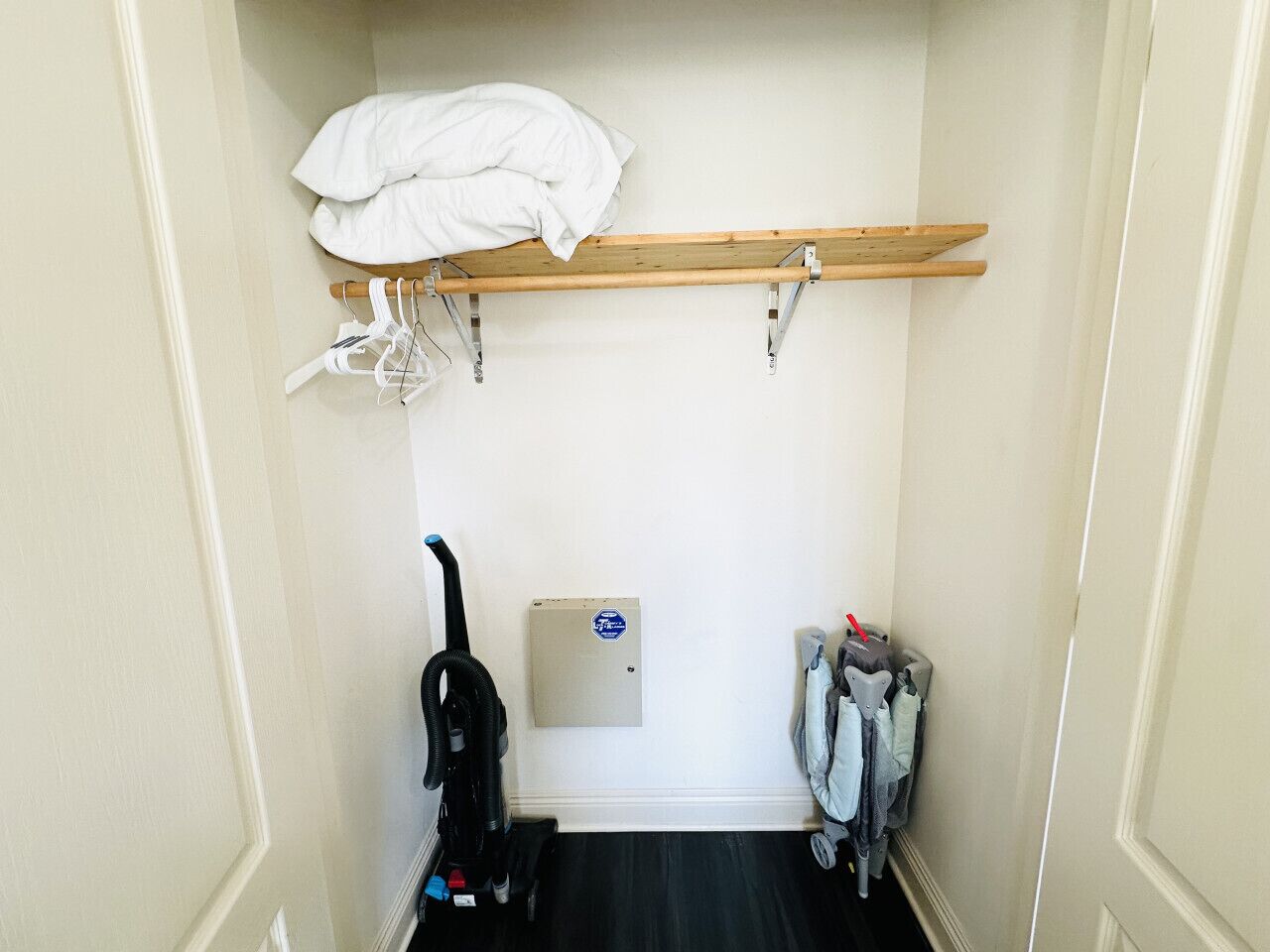 A small, narrow room with a wooden shelf at the top holding a white blanket. A vacuum cleaner is on the left, and a set of baby car seats is on the right.