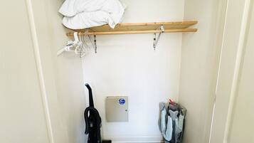 A small, narrow room with a wooden shelf at the top holding a white blanket. A vacuum cleaner is on the left, and a set of baby car seats is on the right.