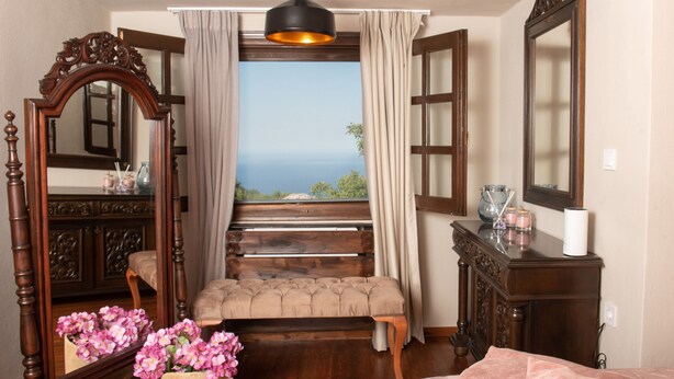 A bedroom with a large window, a wooden bench, a mirror, and a bedside table. - Dandy Villas Tsagkarada | Sky Blue Pansy | Jacuzzi (Tsagkarada)