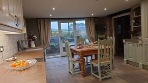 Un coin repas avec une table dressée pour deux personnes, un panier de fruits sur la table et une vue sur l’extérieur par une porte vitrée.