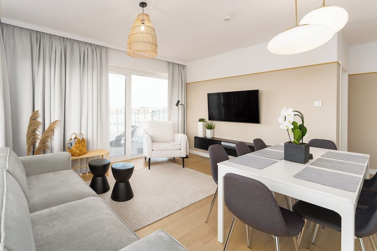 Una sala de estar moderna con un sofá gris, una mesa de comedor con sillas, un televisor montado en la pared y un ventanal con cortinas.