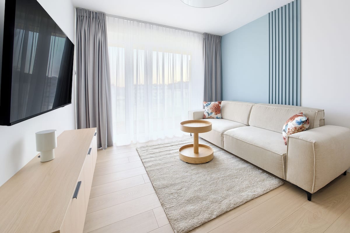 Una sala moderna con sofá, una mesita redonda, televisor y un ventanal grande con cortinas.