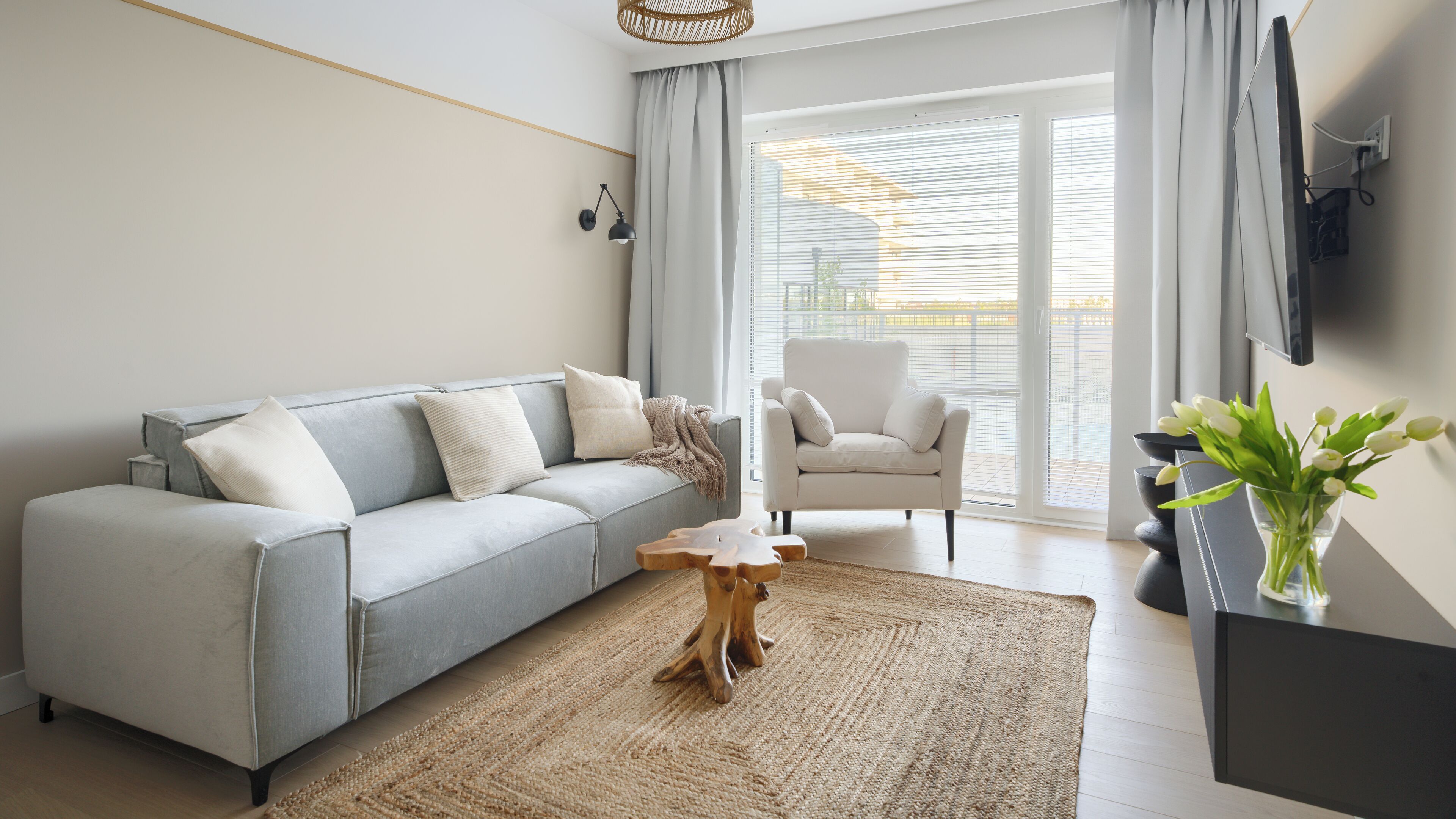 Ein modernes Wohnzimmer mit grauem Sofa, einem Holztisch, einem weißen Sessel und einem großen Fenster mit Jalousien.