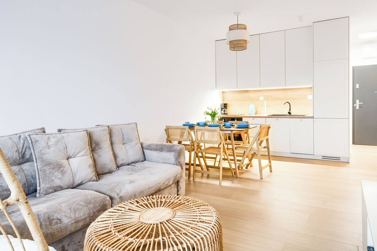 Una sala moderna con un sofá gris, una mesa de comedor de madera y sillas. Hay una zona de cocina con armarios blancos y un refrigerador negro.