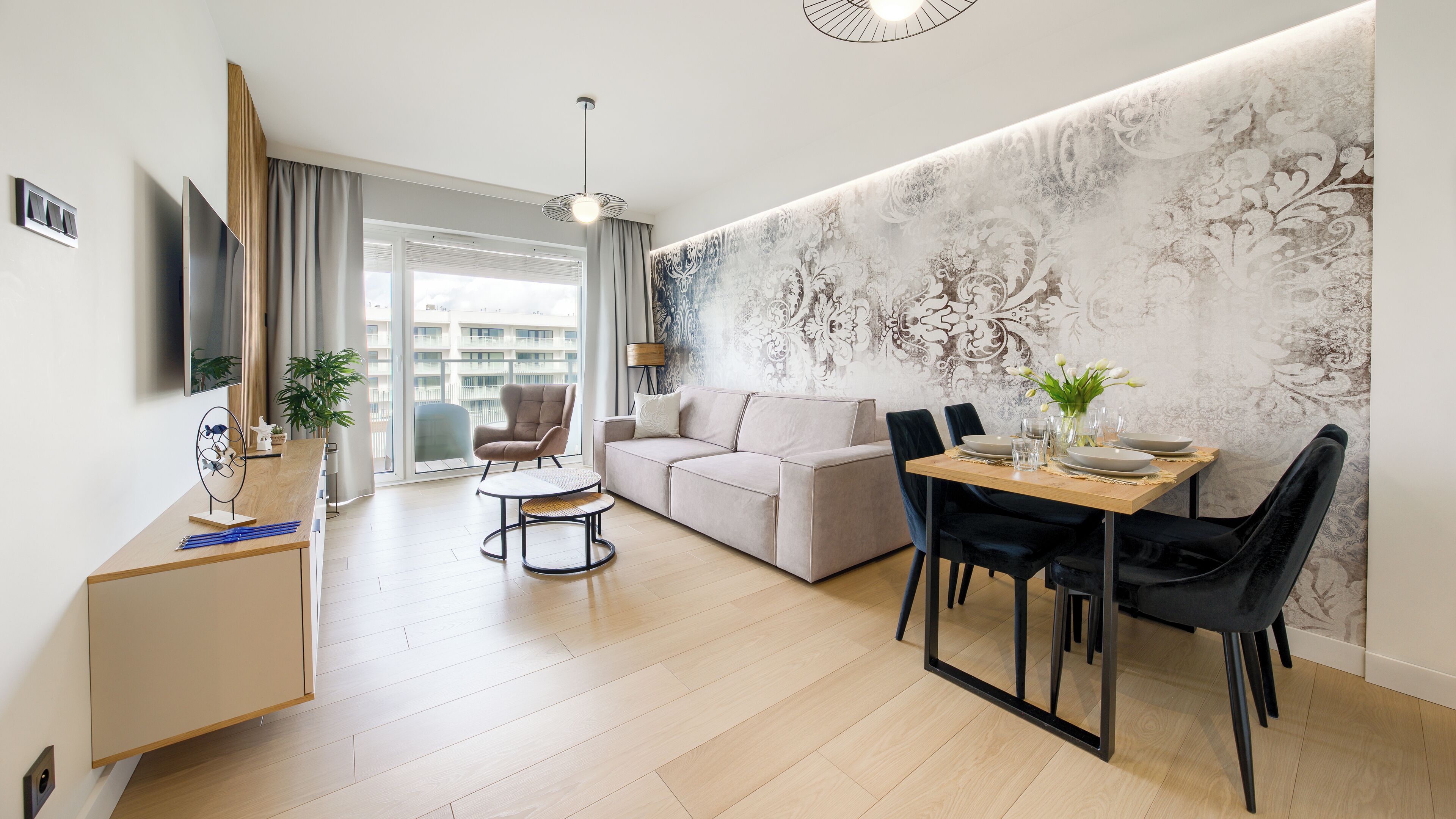 A modern living room with a sofa, dining table, and chairs, a TV, and a large window with a view of buildings.