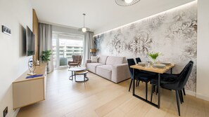 A modern living room with a sofa, dining table, and chairs, a TV, and a large window with a view of buildings. - Apartamenty Sun & Snow Polanki Aqua (Kolobrzeg)