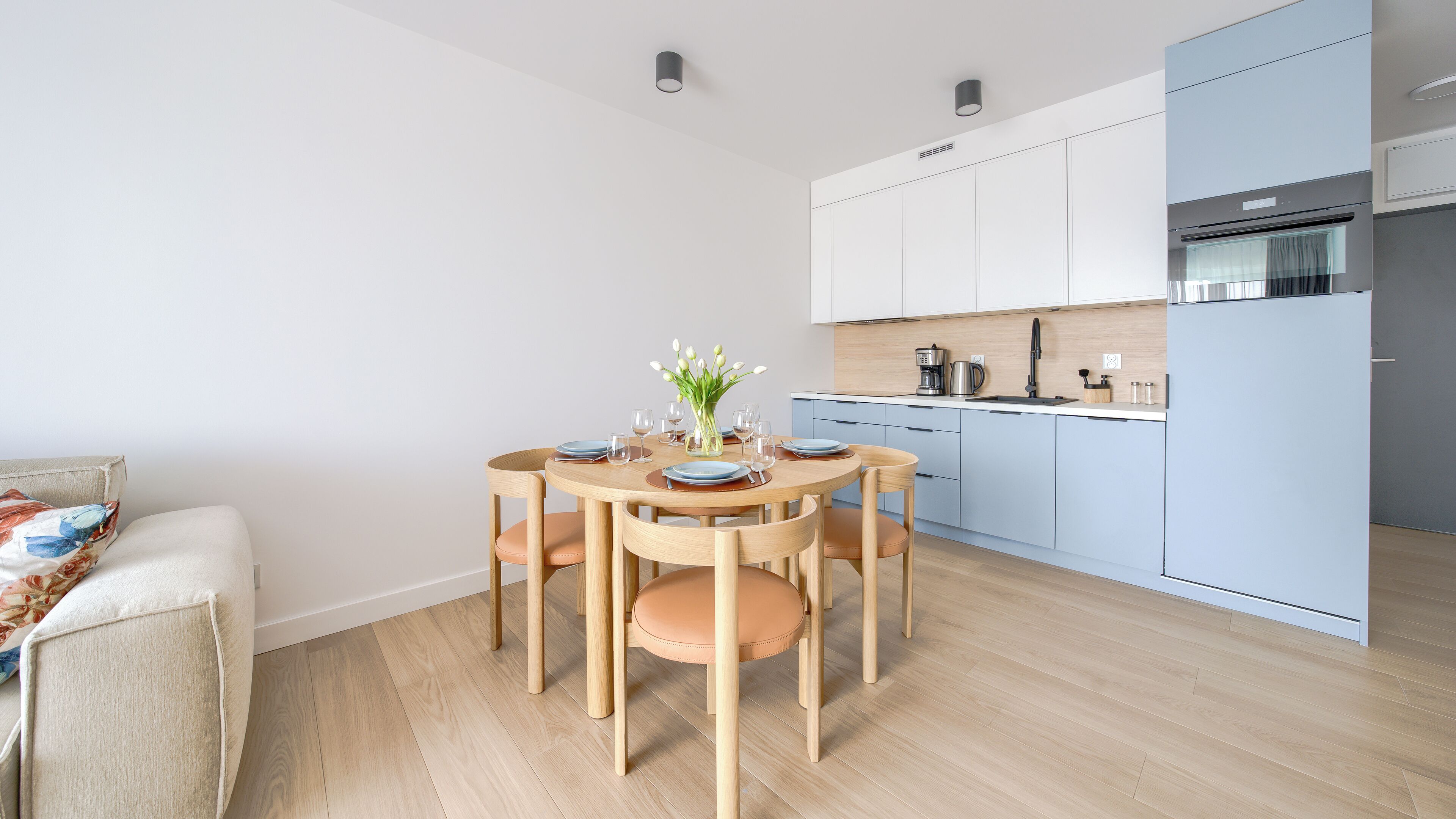 A modern kitchen with light blue cabinets, a round wooden dining table with six chairs, and a small side table with a vase of flowers.