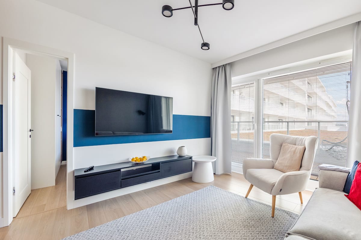 Una sala de estar moderna con un televisor de pantalla plana montado en la pared, un sofá, una silla, una mesita blanca grande y un ventanal con persianas.