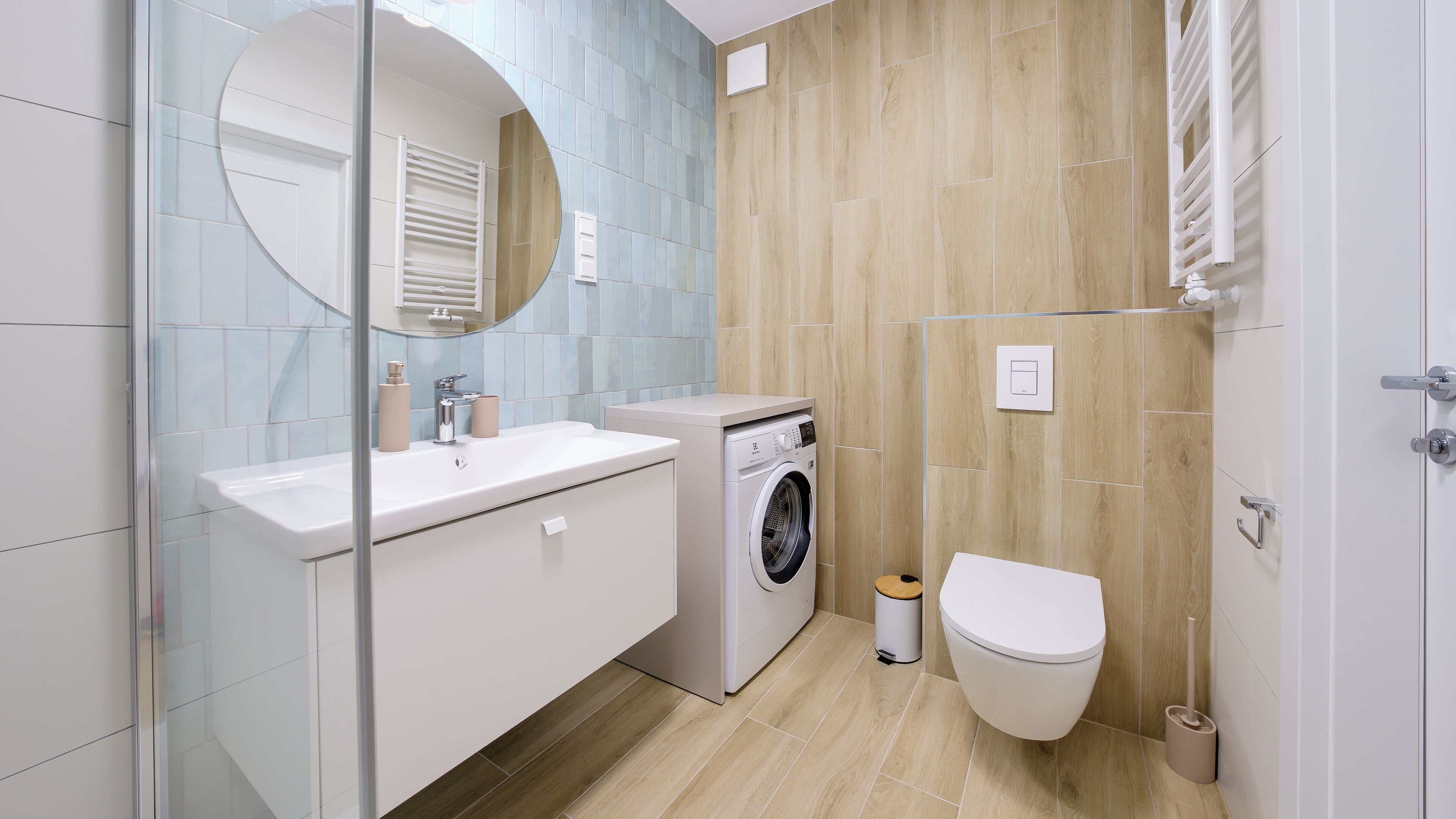 Un baño moderno con cabina de ducha de vidrio, lavadora integrada, inodoro y espejo con marco redondo.