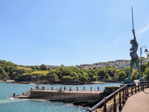Un muelle costero con una estatua, cielo azul despejado y un pueblo en la ladera al fondo.