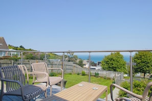 Ein Balkon mit Tisch und Stühlen, Blick auf das Meer und ein umzäunter Garten.