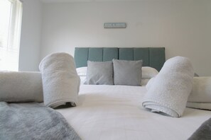 Una cama bien hecha con almohadas y toallas enrolladas, un cabecero verde y un cartel en la pared.