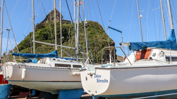 Un puerto deportivo con varios veleros amarrados, una isla destacada al fondo y un edificio con fachada de piedra.