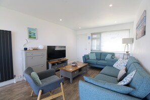 Una sala de estar con televisor, sofá gris, sillón azul, mesa de centro de madera y una lámpara.