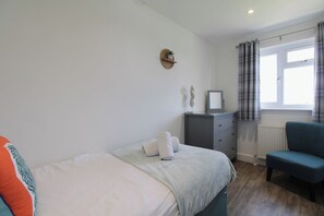 Um quarto com cama, cômoda, cadeira, janela com cortinas e um enfeite na parede.