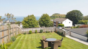 Un jardin à l’arrière d’une maison, avec une pelouse, une table ronde entourée de chaises, une clôture en bois et, au loin, une vue sur la mer et les collines.