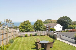 A residential backyard with a lawn, a round table with chairs, a wooden fence, and a view of the sea and hills in the background.