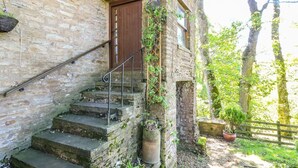 Kivestä rakennettu portaikko johtaa puiselle ovelle, ja sen ympärillä on vehreää kasvillisuutta sekä maalaishenkinen kivimuuri.
