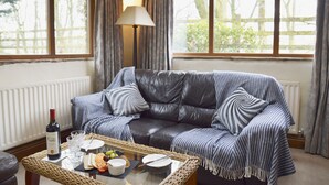 A cozy living room with a leather sofa, a glass coffee table with snacks and drinks, and large windows with curtains. - Bothy (Alston)