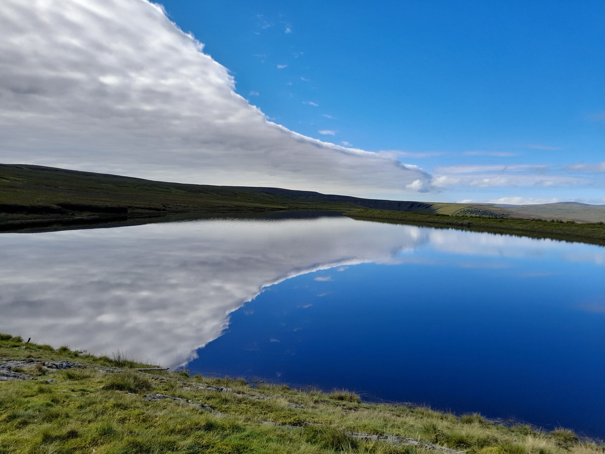 A calm lake with a clear reflection of the sky and surrounding hills.