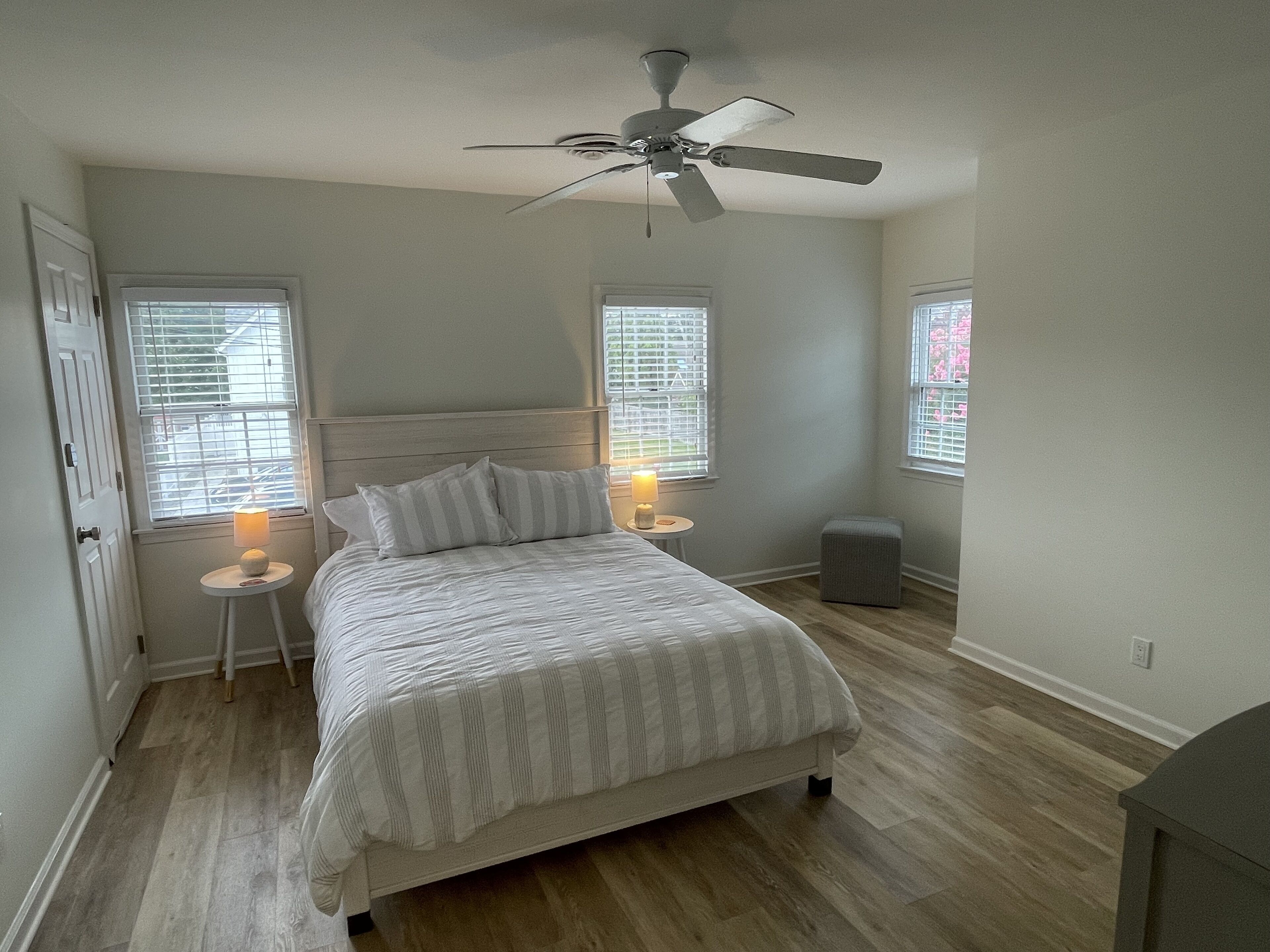 Une chambre à coucher comprenant un lit, deux tables de chevet avec des lampes, un ventilateur de plafond et deux fenêtres équipées de stores.