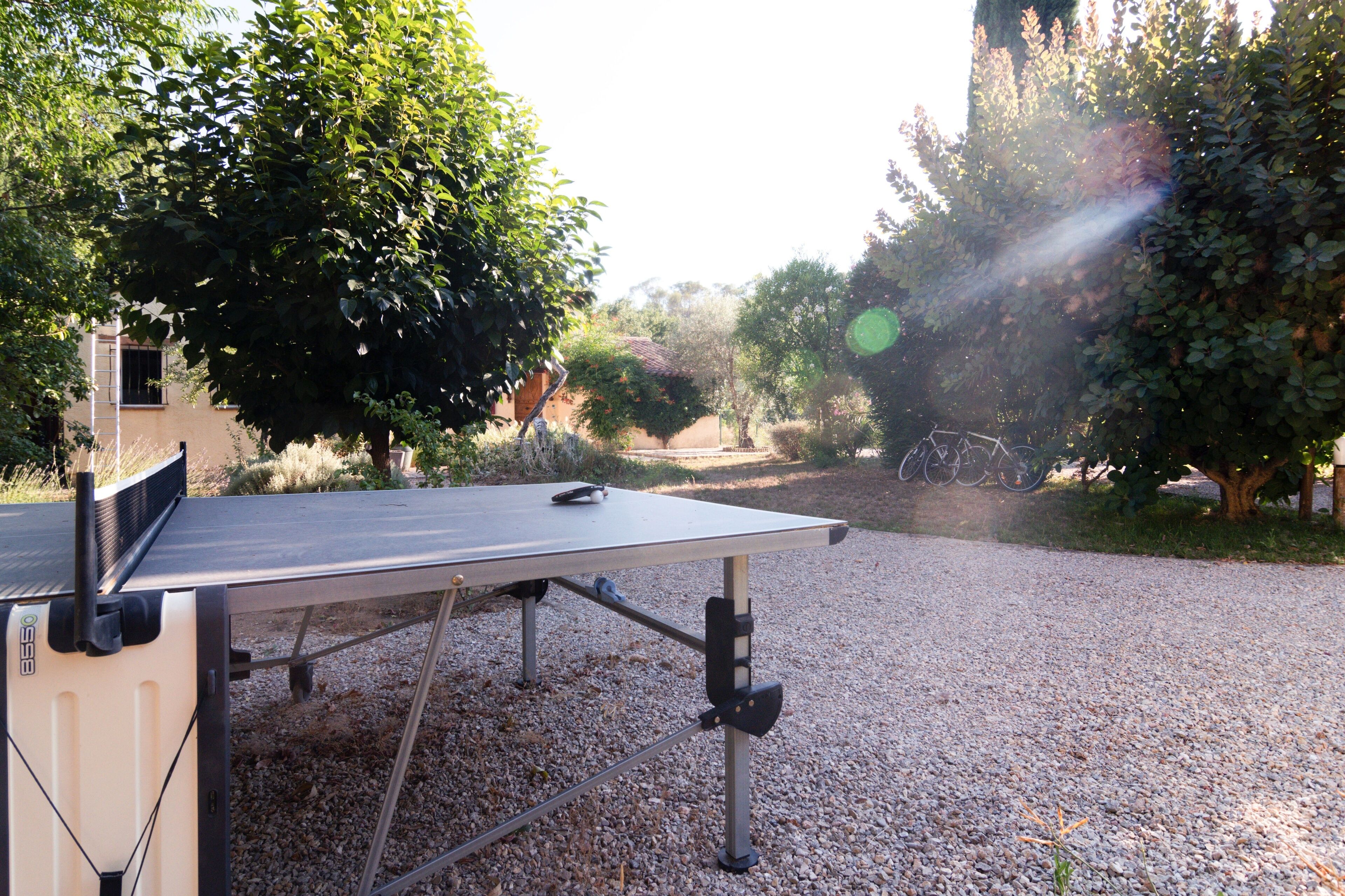 A ping pong table in an outdoor setting with gravel ground, surrounded by trees and a building in the background.