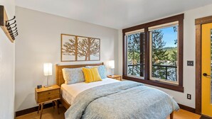 Une chambre à coucher comprenant un lit, deux lampes de chevet, une table de nuit, une fenêtre donnant sur des arbres et une porte.
