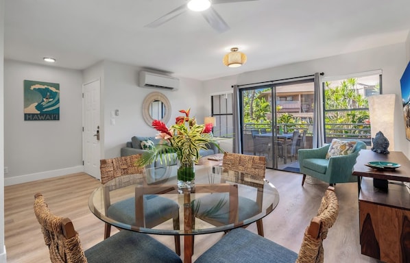 A modern living room with a glass dining table, wicker chairs, a sofa, and a view of a tropical garden through large sliding doors.