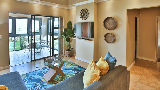 A spacious living room with a grey sofa, a glass coffee table, and a large plant. There is a sliding glass door leading to a patio with outdoor furniture.