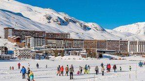Un complejo de esquí con varios edificios, incluyendo un gran hotel de varios pisos y balcones, rodeado de montañas nevadas.