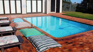 Une piscine rectangulaire bordée de carreaux, entourée d’une terrasse en briques. On y trouve des chaises longues avec des coussins et un oreiller à rayures.
