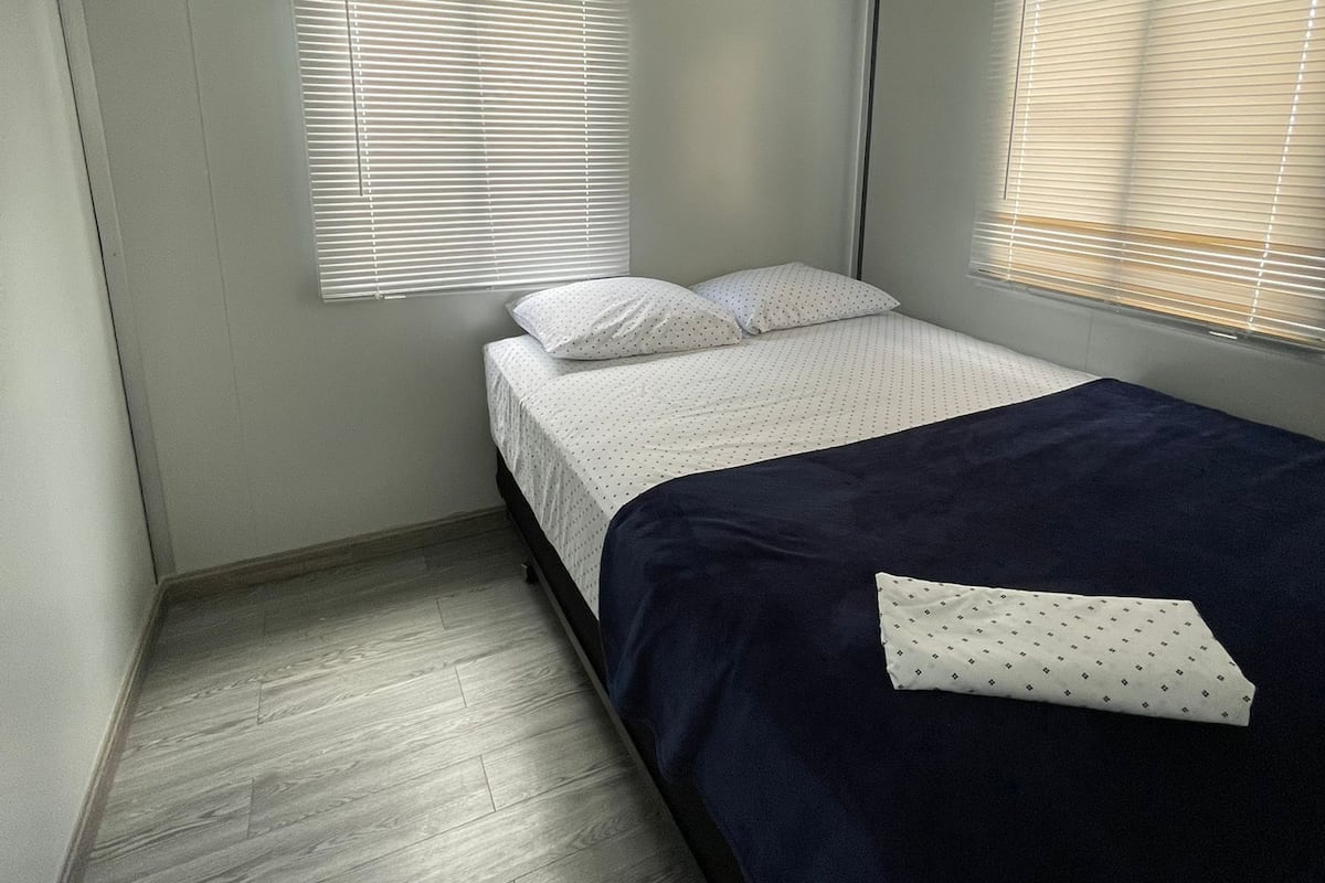 Ein kleines, helles Schlafzimmer mit einem Einzelbett, zwei Fenstern mit Jalousien, einer Deckenleuchte und einem Holzboden.