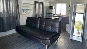 A modern living room with a black leather sofa, a kitchen area with a refrigerator and microwave, and a sliding glass door leading to a balcony. - Las Casitas de Village (Carolina)