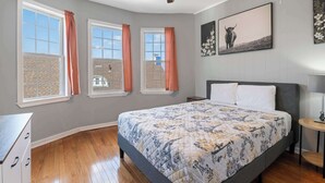 A bedroom with a large bed, a ceiling fan, three windows with curtains, and artwork on the walls. - Rustic Paradise - Steps to Beach! (Atlantic City)