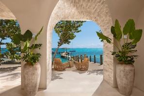 A view through an arched doorway to a pool area with wicker chairs and potted plants, overlooking a clear blue sea and a dock. - Impression Isla Mujeres by Secrets – Adults only – All Inclusive (Isla Mujeres)