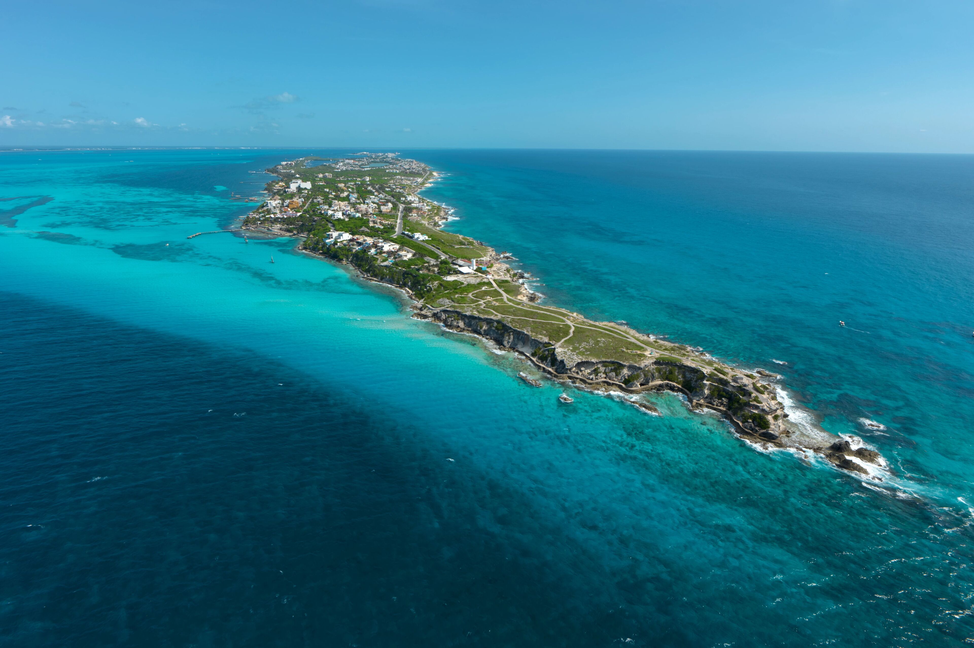 Vue aérienne d’une île côtière aux eaux turquoise et limpides, avec une étroite bande de terre parsemée de bâtiments et un littoral accidenté.