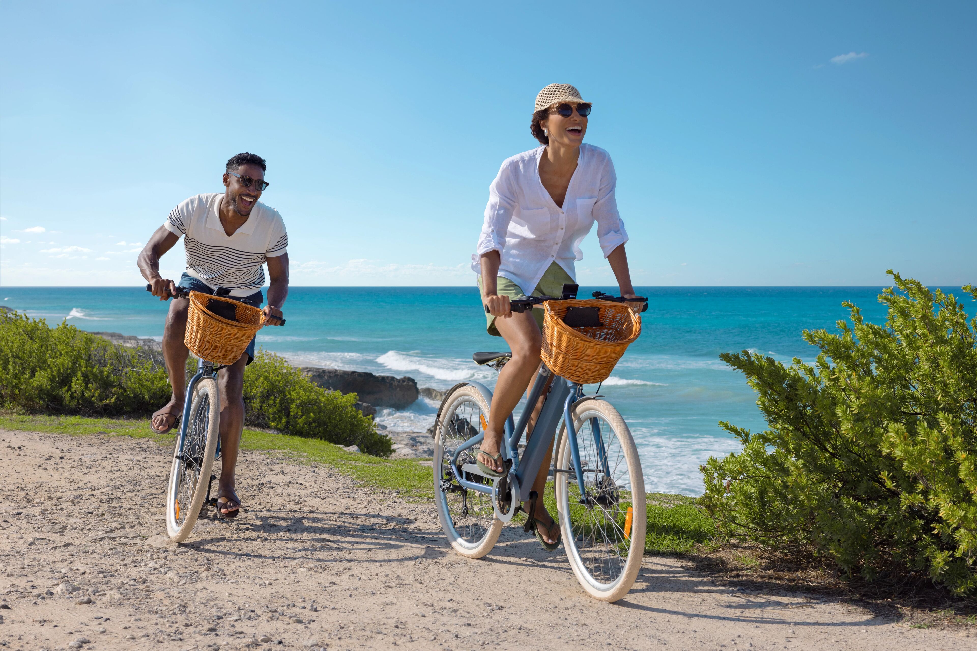 Dos personas andan en bicicleta por un sendero arenoso con vista al océano.